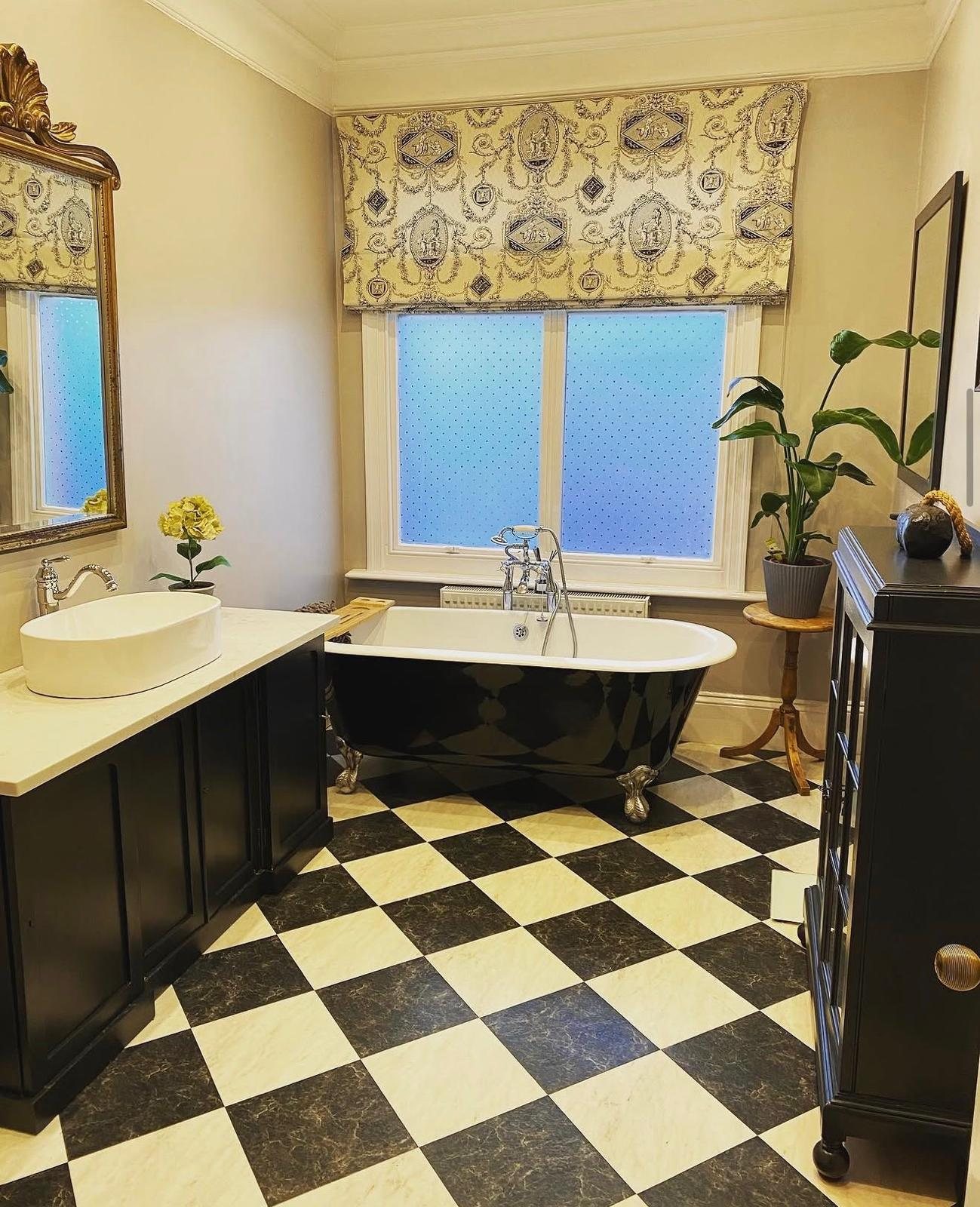 Bathroom with a black roll-top bath, chequerboard floor and classical print roman blind